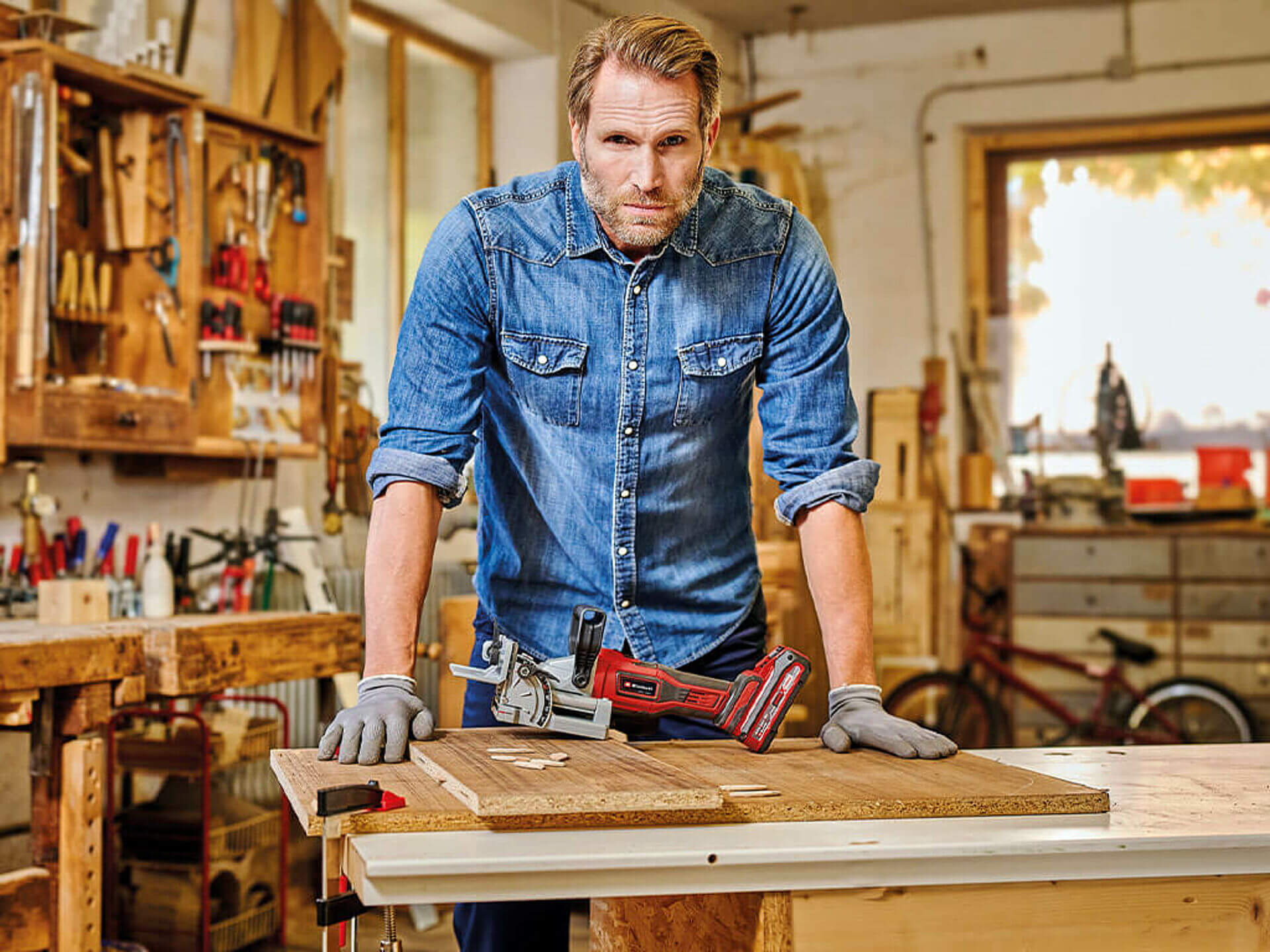 A man is standing in a workshop behind a workbench on which the Einhell cordless biscuit jointer is lying.