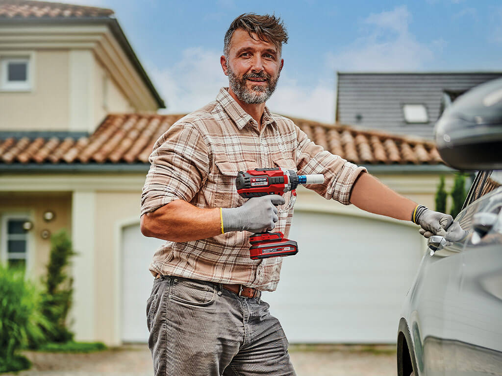 A man is standing next to his car holding an Einhell cordless impact wrench.